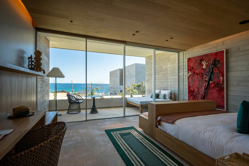 Modern bedroom with large windows, ocean view, and minimalistic decor. Features a wooden ceiling, bed, lounge chair, and a vibrant red artwork on the wall.