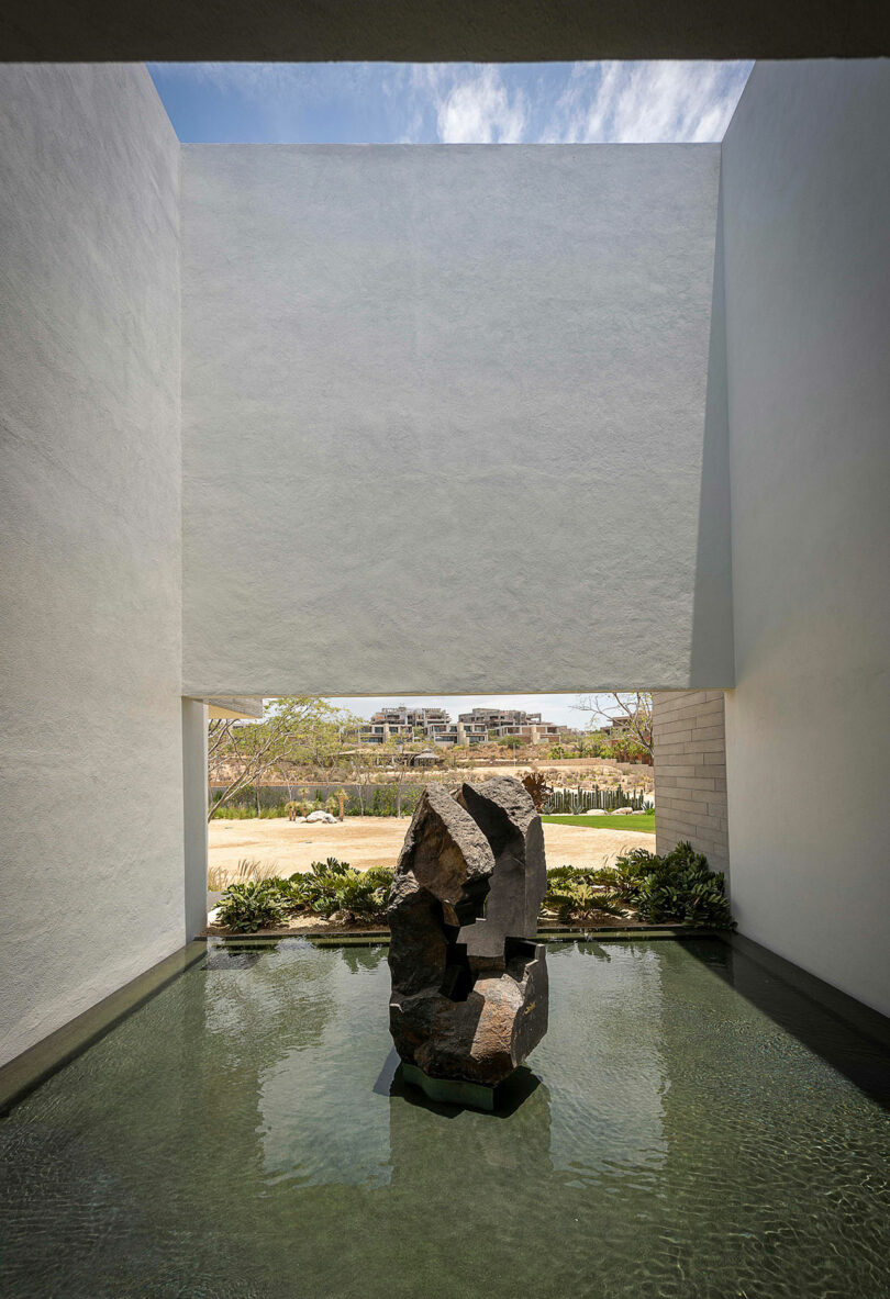 Sculpture of stacked stones in a shallow reflecting pool with white walls and a view of the outdoors.