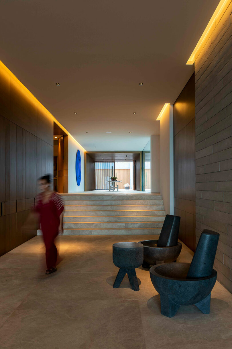 A person walks through a contemporary hallway with soft lighting, stone floors, black chairs, and a stairway leading to a room with a circular wall art and a visible window at the back.