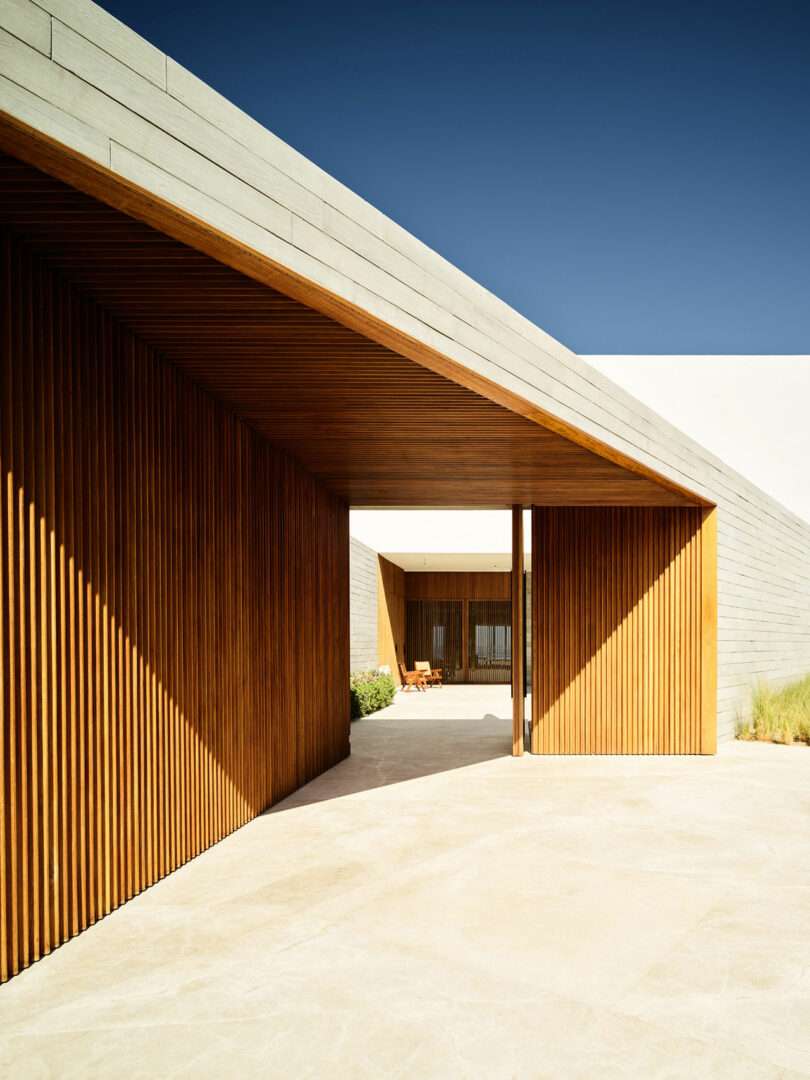 Modern architectural structure with wooden slats, a light stone pathway, and a clear blue sky. A chair is visible in the shaded area.