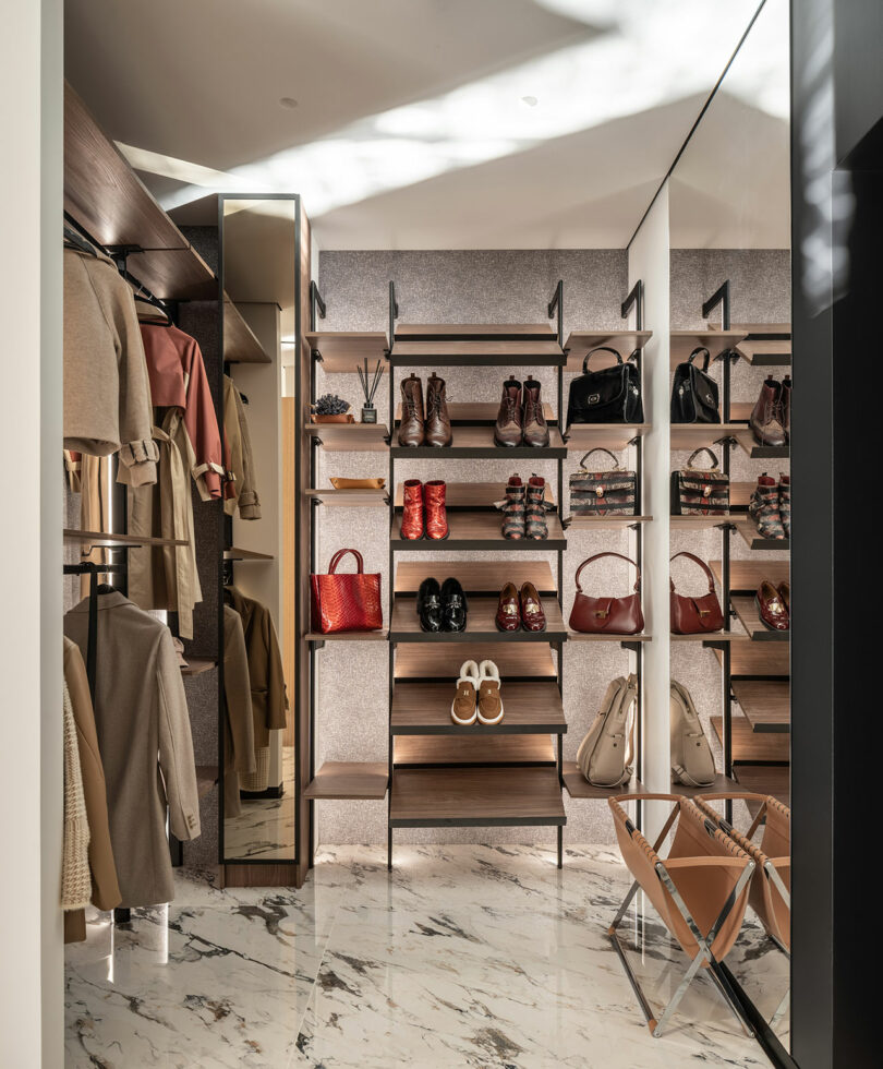 Luxurious walk-in closet with marble floor, featuring shelves of shoes and bags, clothing racks with coats and dresses, a full-length mirror, and a folded chair.