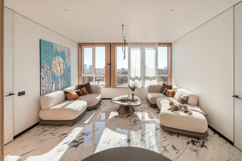 Modern living room with light beige sofas, a round coffee table, and a large abstract painting. Marble floor, large windows with sheer curtains, and a dog resting on one of the sofas.