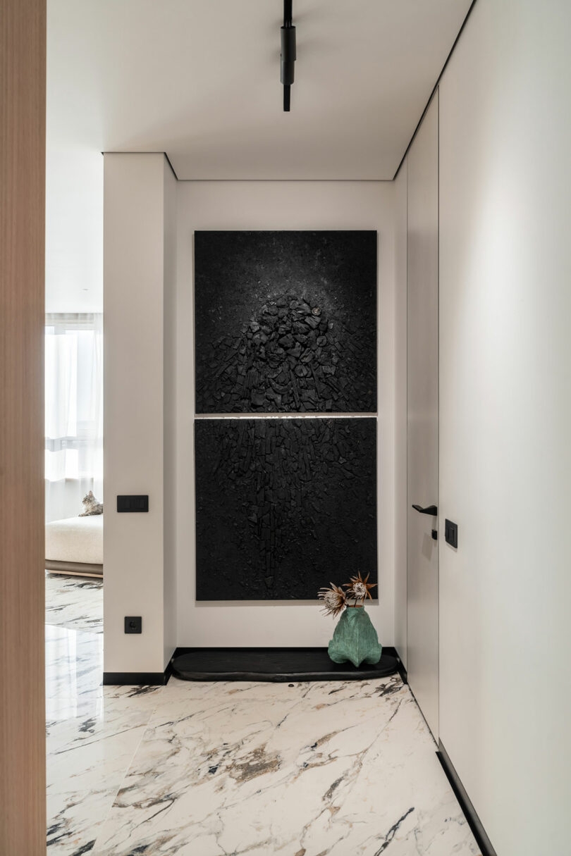 Narrow hallway with marble floor, black textured wall art, and a turquoise vase with dried branches. Bright natural light enters from a room at the end of the hall.