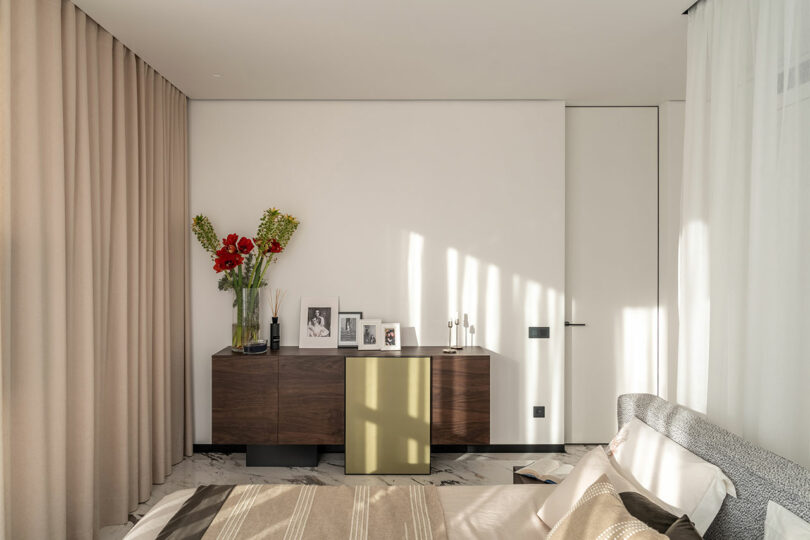 A bedroom corner with a wooden sideboard holding framed photos and a vase of red flowers. Beige curtains, a bed, and a door are visible. Light creates shadows on the wall.