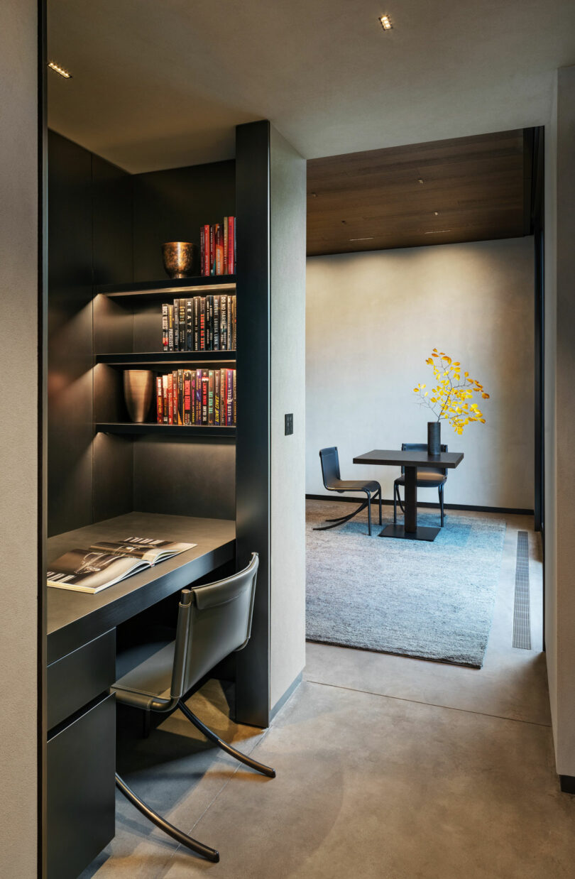 Modern interior with a study nook featuring a desk, chair, and bookshelf. In the background, a dining table with chairs and a vase with yellow flowers on a rug.