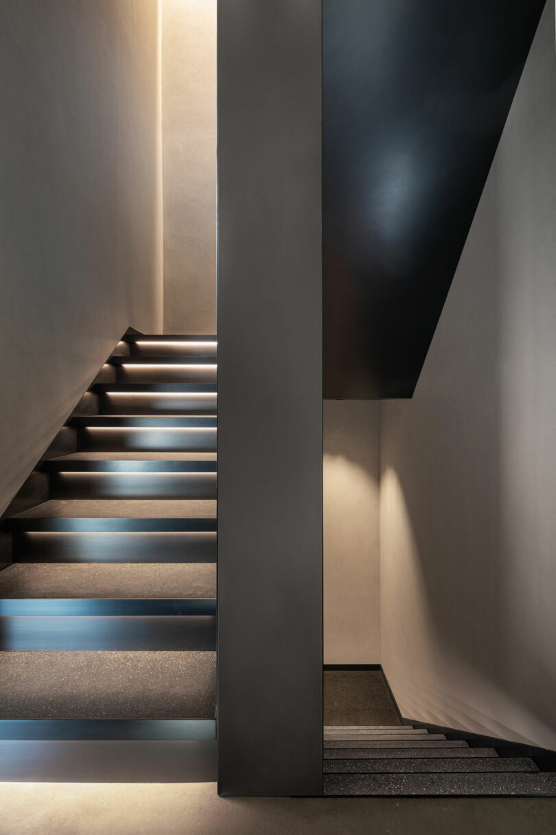 Minimalist interior with two sets of stairs divided by a wall; one ascending on the left and the other descending on the right, both with soft lighting.