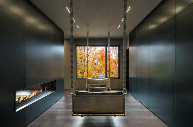 Modern room with dark walls, a suspended sofa facing a large window with autumn trees visible outside, and a linear fireplace on the left wall.