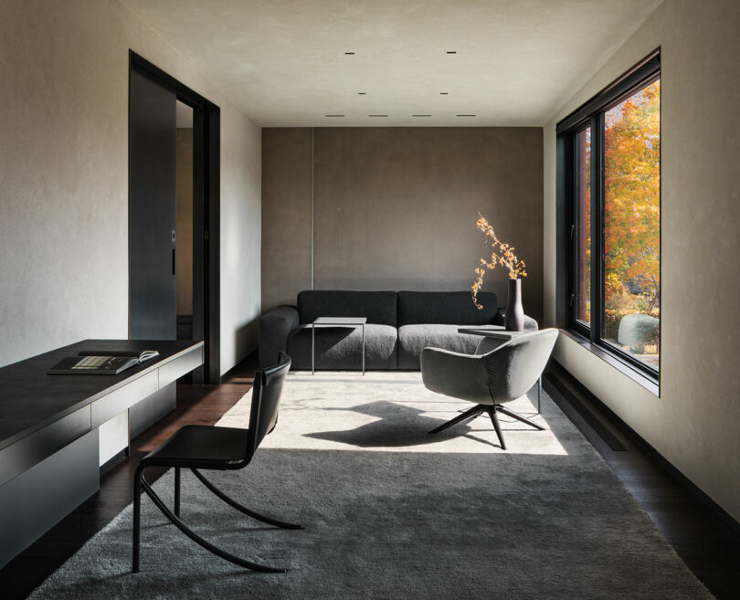 Minimalist living room with gray sectional sofa, lounge chair, and large window showcasing autumn foliage. Neutral tones dominate with a dark rug and simple decor.