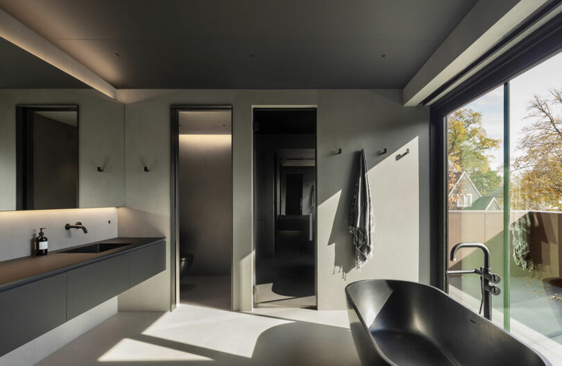 Modern bathroom with matte black fixtures, minimalist design, large mirror, towel on a hook, and a freestanding tub next to a floor-to-ceiling window with a view of trees.