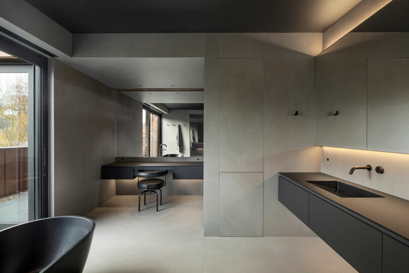 Modern minimalist bathroom with gray walls, large mirror, built-in cabinets, freestanding black bathtub, and wall-mounted sink with soft lighting.