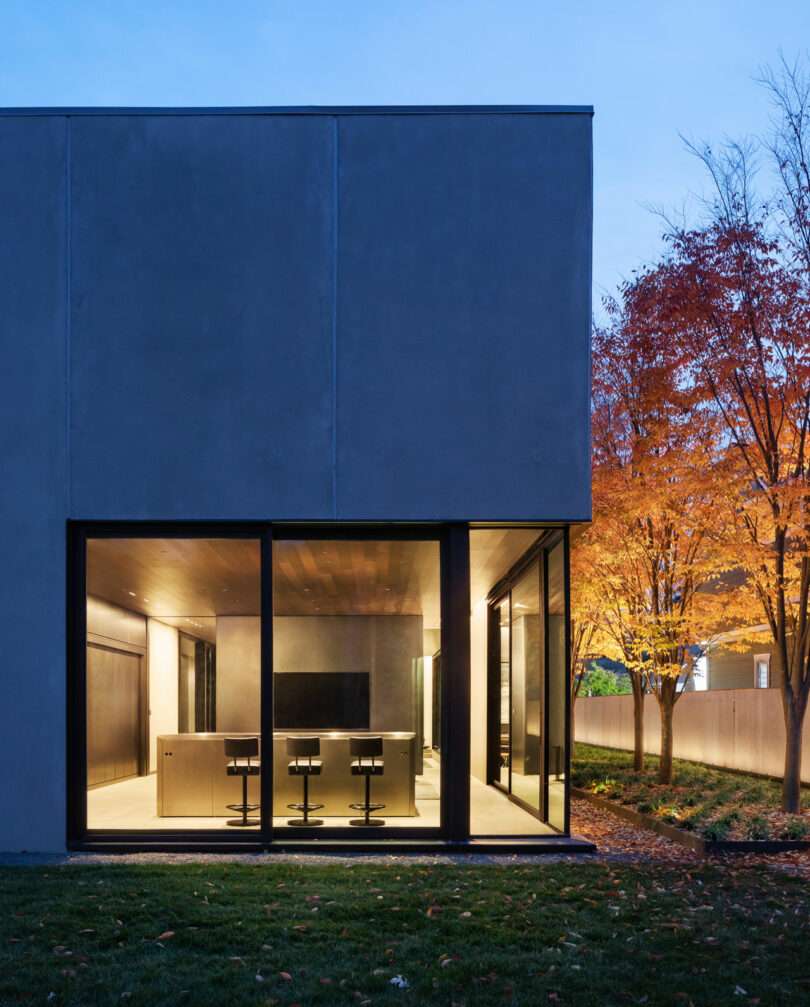 Modern building with large glass windows revealing a bright interior with chairs and a table. It's adjacent to a lawn and a tree with autumn-colored leaves, set against a twilight sky.