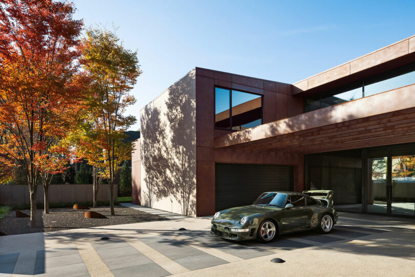 Modern house with large windows and a brown facade, featuring an olive-green sports car parked on the driveway. Autumn trees with orange leaves are on the left.