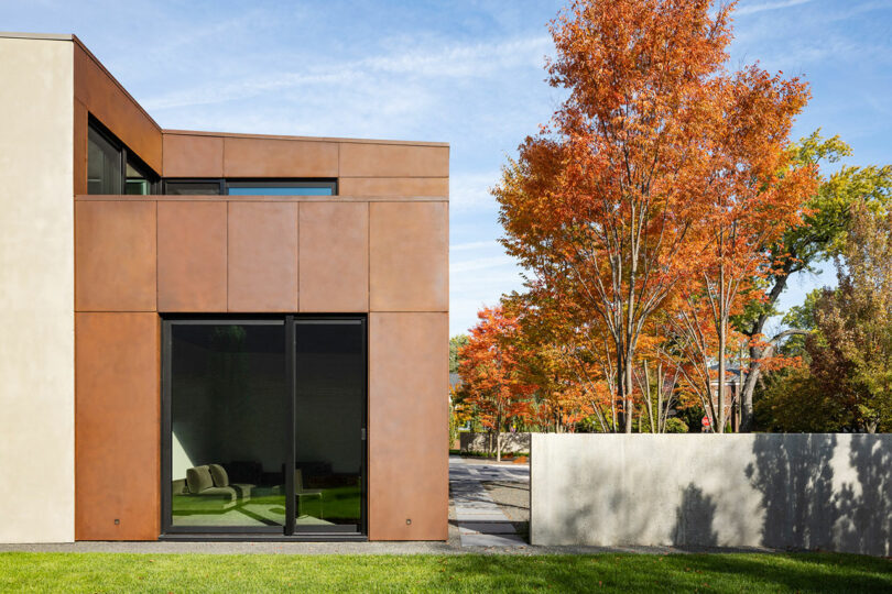 Modern house exterior with large glass windows, surrounded by a green lawn and vibrant autumn trees.