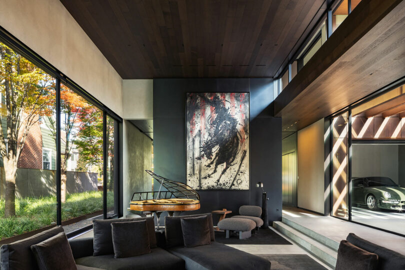 Modern living room with large windows, dark furniture, abstract artwork, and a decorative piano. Glass wall reveals a parked car in the adjacent area.