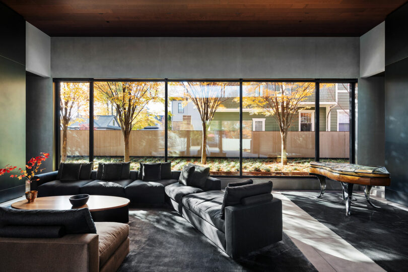 Modern living room with large windows, dark grey sofas, a round table with red flowers, and a piano. Sunlight streams in illuminating autumn trees outside.