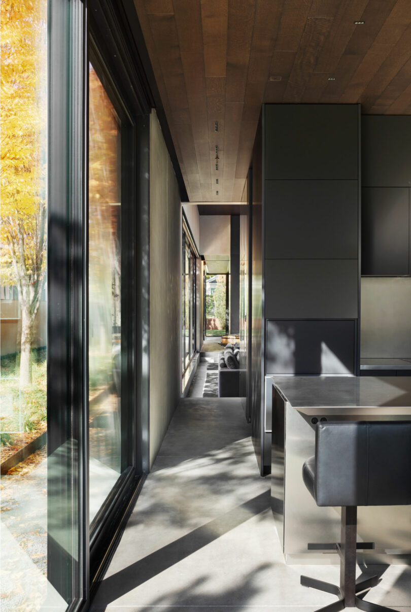 Modern interior hallway with large windows on the left, black cabinetry on the right, and a sleek black counter with chairs in the foreground. Sunlight streams in, casting shadows on the floor.