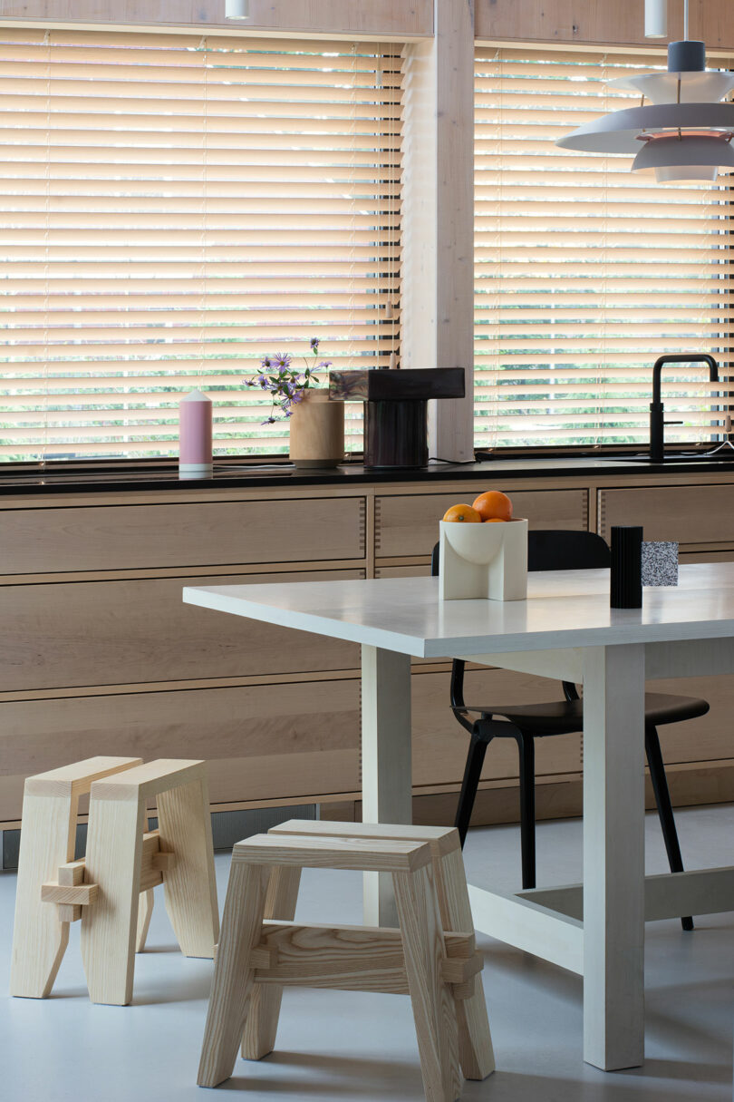 Modern kitchen with a white table, two wooden stools, a black chair, and a countertop with a fruit bowl. Windows with wooden blinds allow natural light in.