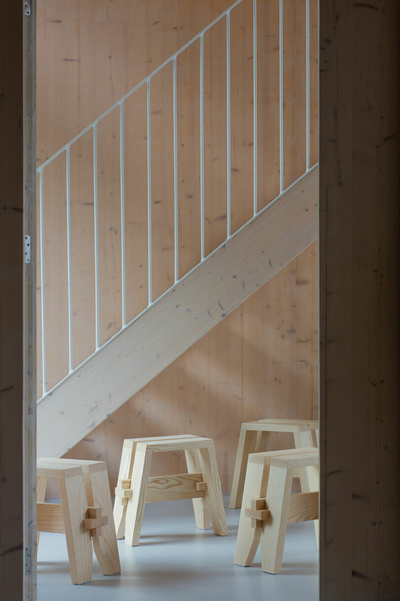 Wooden stools are arranged on a floor beneath a wooden staircase with metal railings.