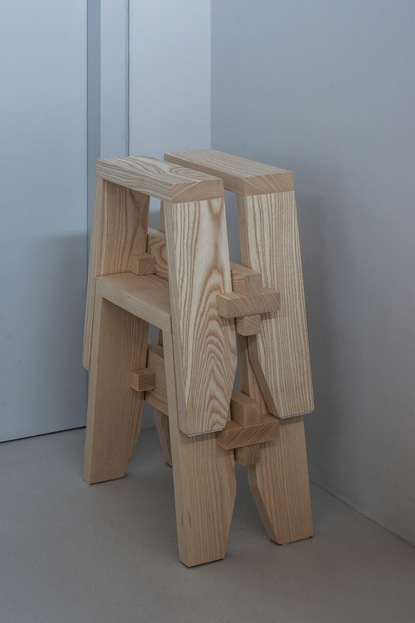 Two interlocking wooden stools with a simple, geometric design are stacked against a light-colored wall.