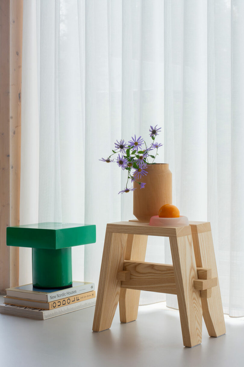 A wooden stool holds a vase with purple flowers and an orange fruit. Nearby, a stack of books supports a green geometric sculpture. Sheer white curtains hang in the background.