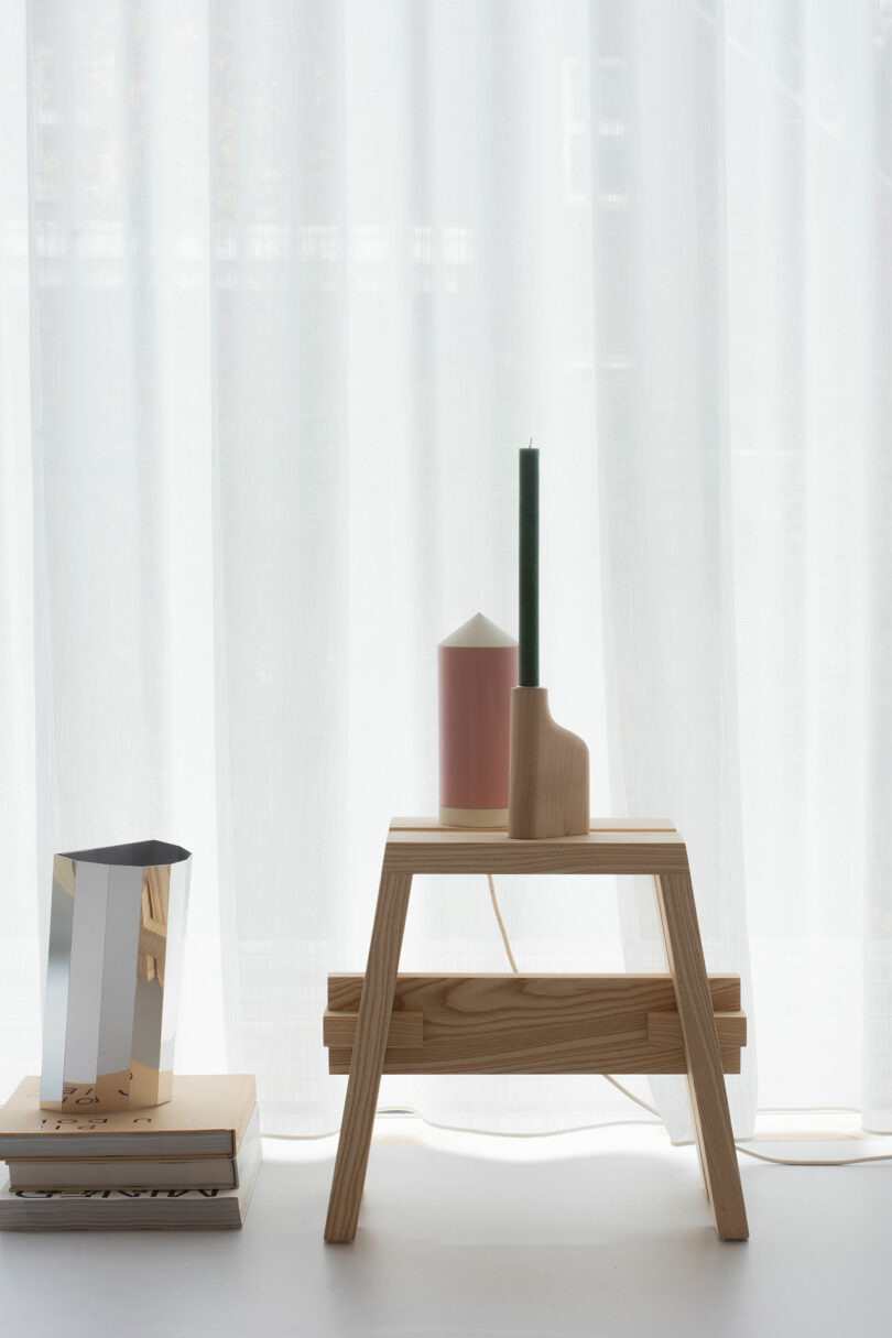 A minimalist wooden table with a green candle and a pink container, beside a silver pitcher on stacked books, in front of a sheer white curtain.