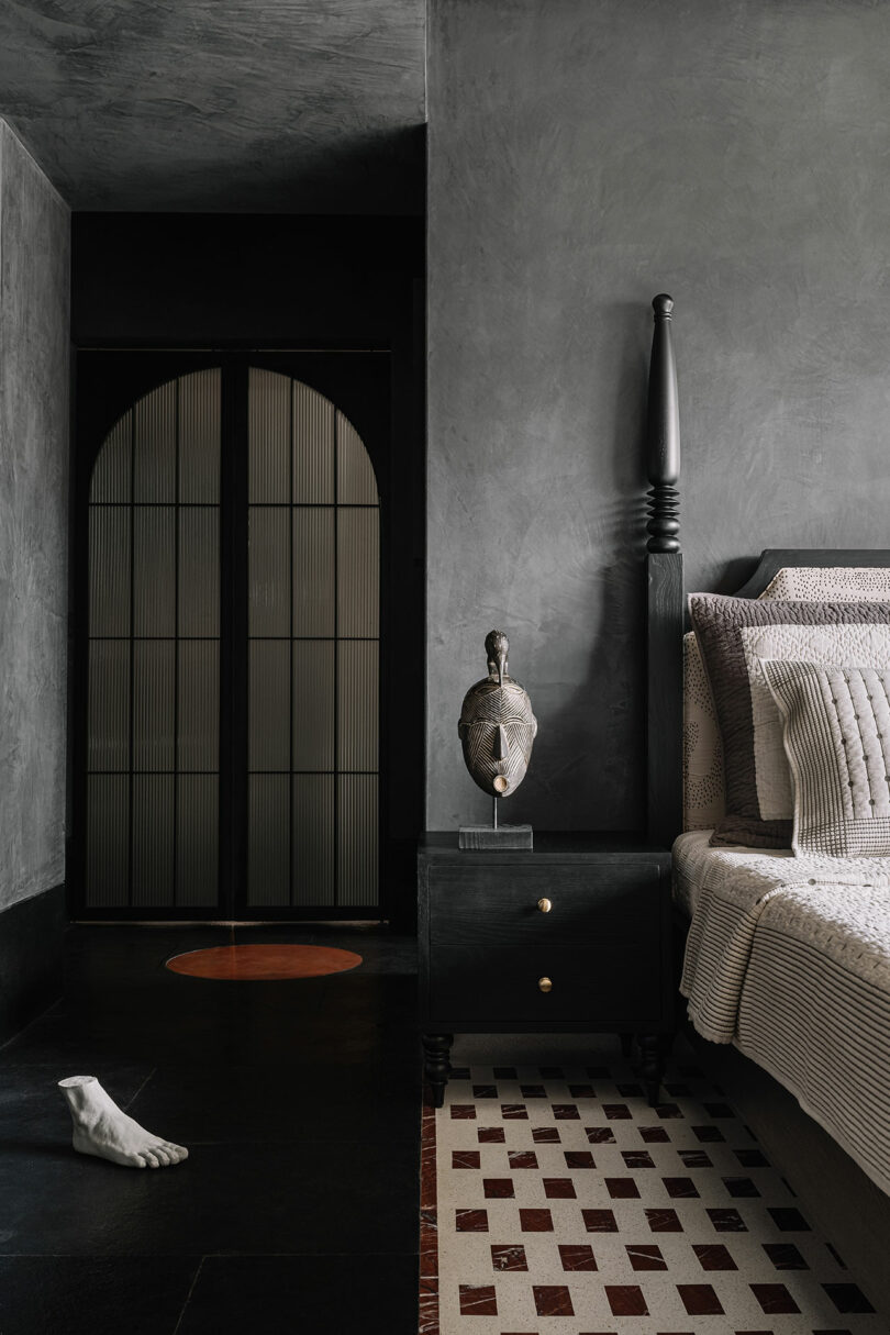 A dimly lit bedroom designed by Studio Nishita Kamdar showcases a dark gray color scheme, featuring a sculpture on a black nightstand, a large bed with textured bedding, a checkered floor, and an arched door in the background.