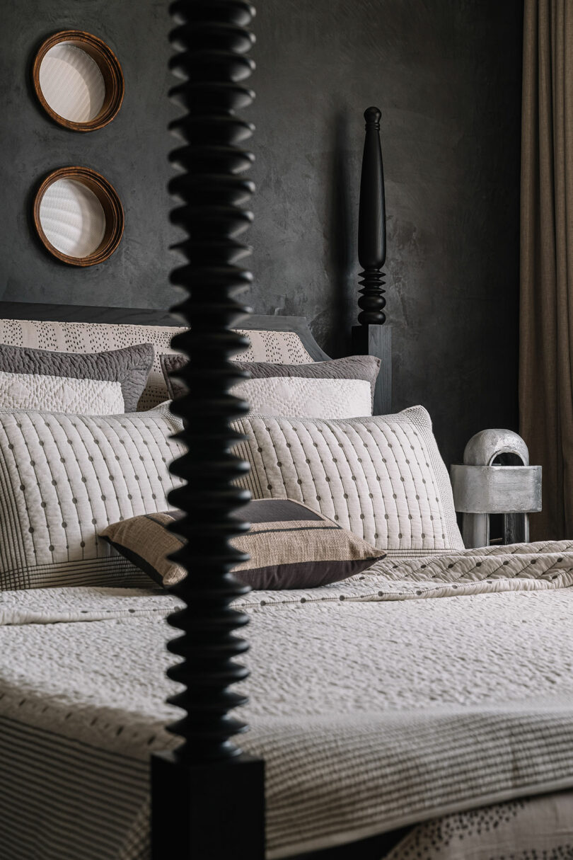 A modern bedroom designed by Studio Nishita Kamdar features a four-poster bed, textured bedding, decorative pillows, a side table, and two wall-mounted round baskets. The dark wall beautifully complements the room's neutral tones.