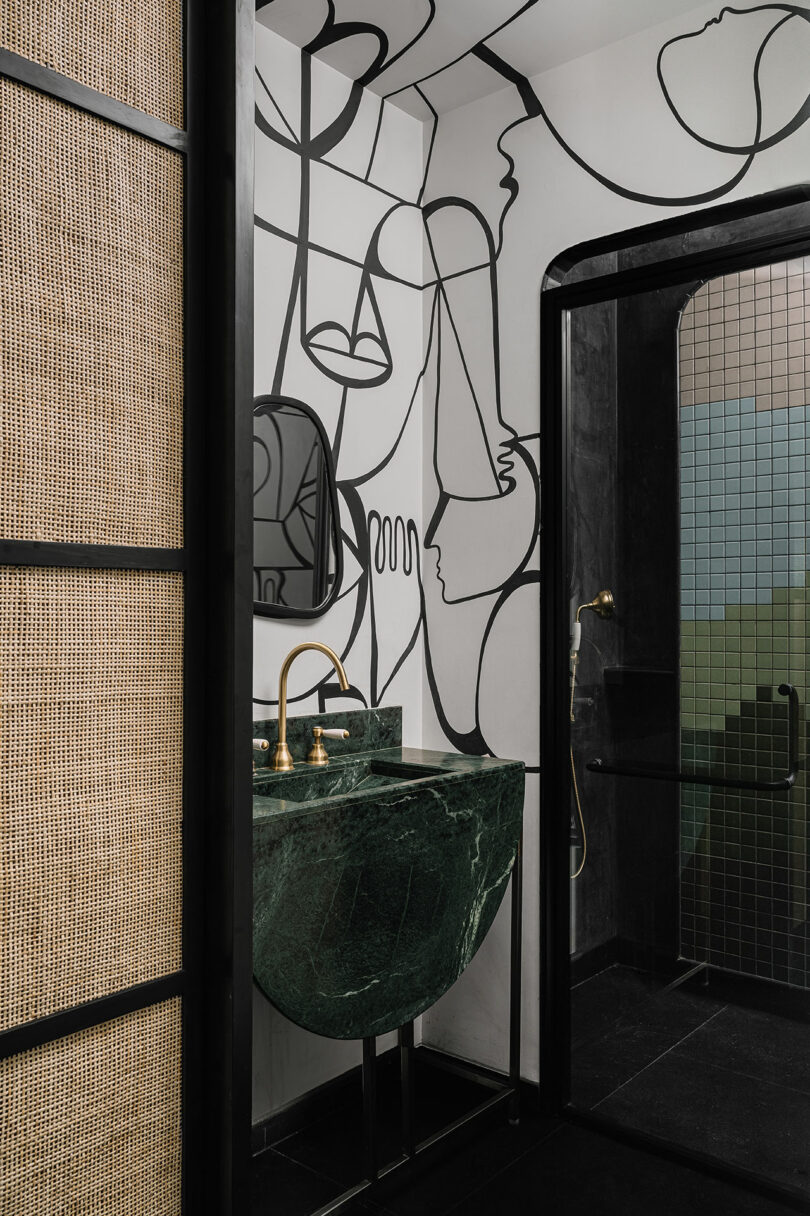 Modern bathroom featuring a green marble sink, gold faucet, and artistic wall mural designed by Studio Nishita Kamdar. The glass-tiled shower area adds a touch of elegance.