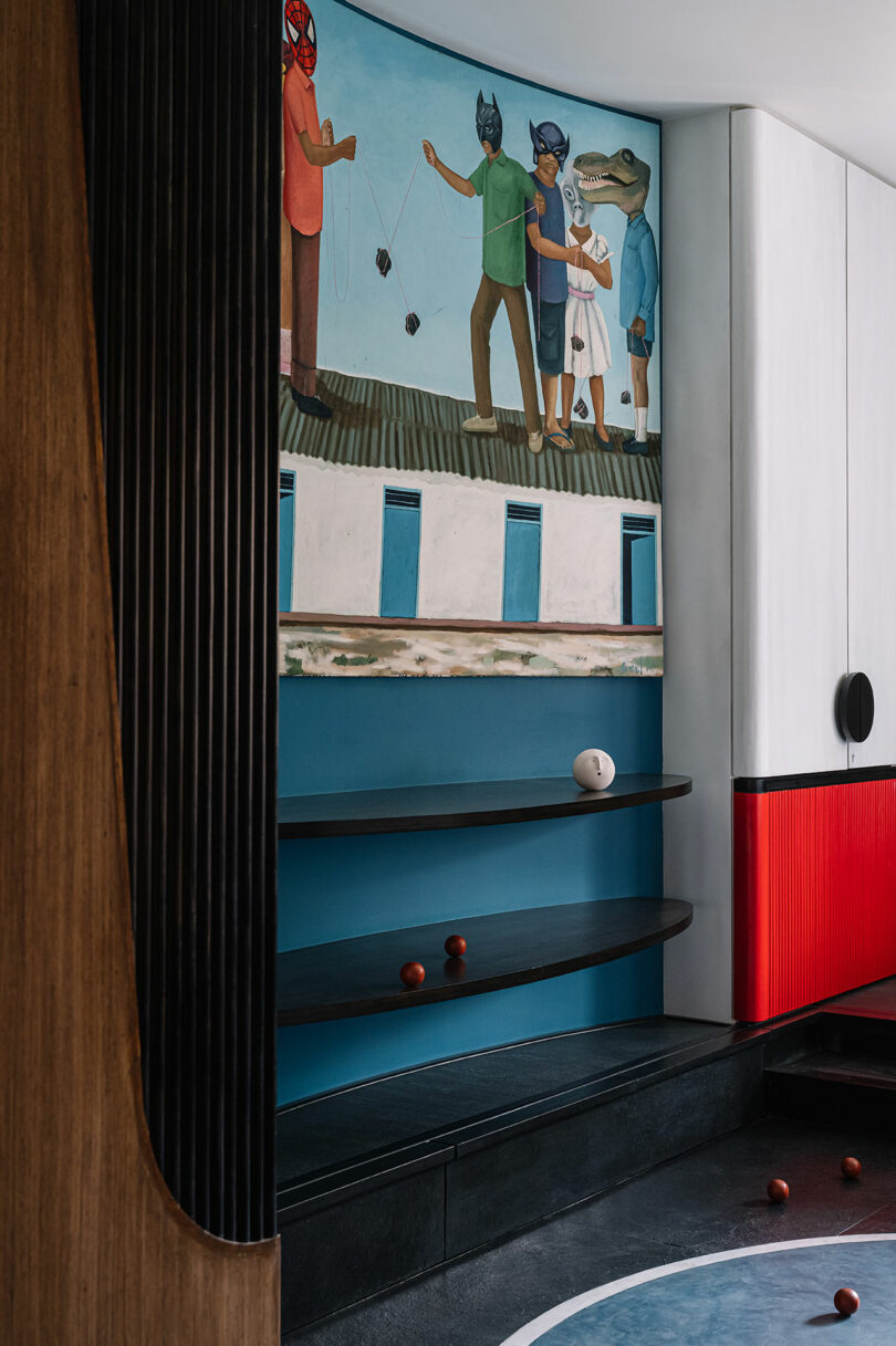 A room designed by Studio Nishita Kamdar features a vibrant mural of people and animals, complemented by curved wooden and red paneling. A blue shelf with scattered red and white balls adds a playful touch.