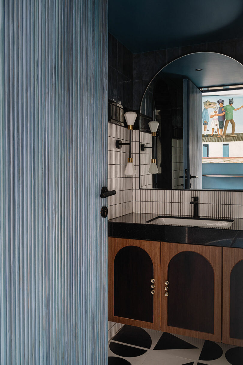 Designed by Studio Nishita Kamdar, the bathroom features a blue ribbed wall, an arched mirror, and a black sink perched on a wooden cabinet. Geometric floor tiles add to the charm, while wall art is elegantly captured in the reflection.