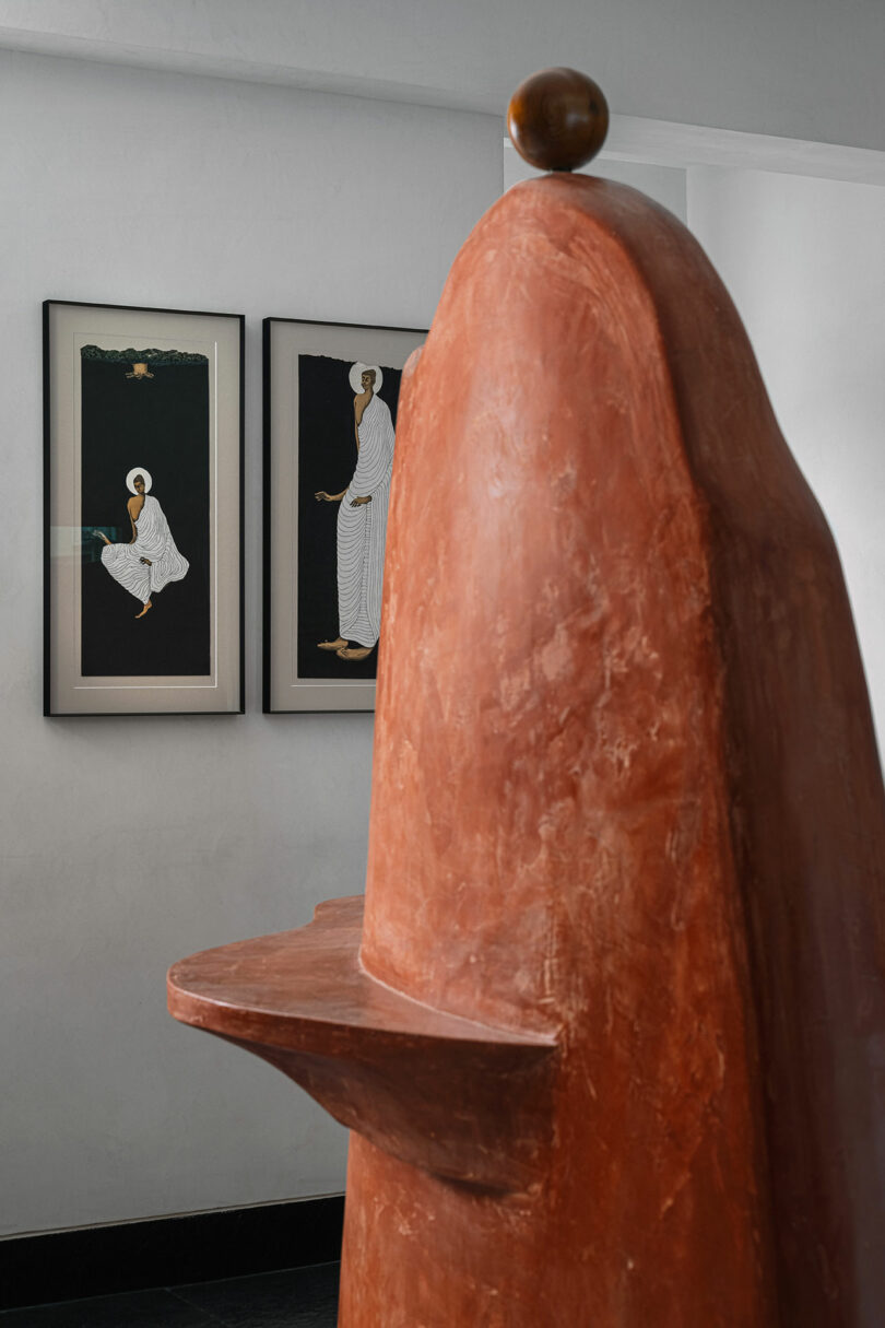 A large, abstract terracotta sculpture from Studio Nishita Kamdar stands in the foreground, with two framed prints depicting figures in white robes on a black background hanging on the wall behind it.