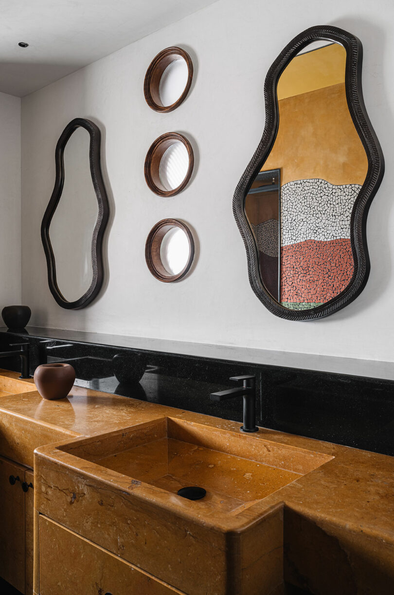 This bathroom, designed by Studio Nishita Kamdar, features two uniquely shaped mirrors, three circular mirrors, and dual brown stone sinks with sleek black faucets.