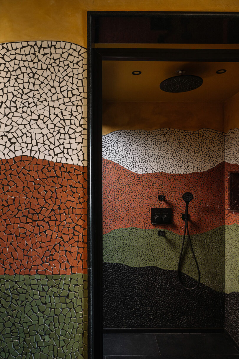 A shower area designed by Studio Nishita Kamdar showcases a mosaic tile pattern in earthy tones, with curving bands of black, green, red, and white. A rainfall showerhead and controls are elegantly visible on the wall.