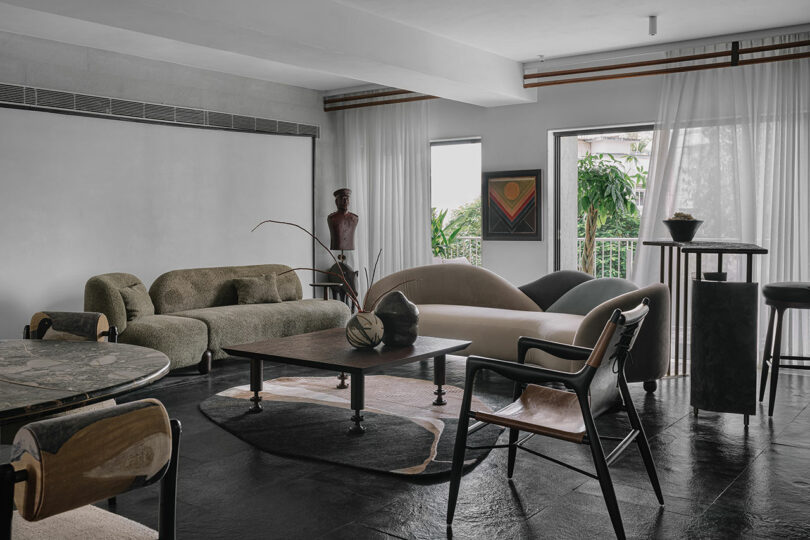 A modern living room designed by Studio Nishita Kamdar features contemporary furniture, including sofas, chairs, and a coffee table. Large windows with sheer curtains allow natural light to flood in. A sculpture and framed artwork enhance the space's elegance.