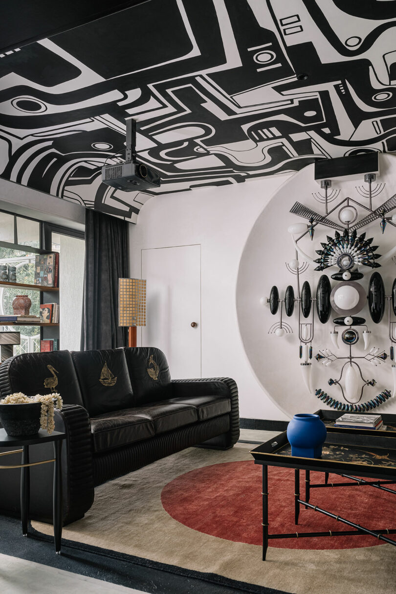 A modern room designed by Studio Nishita Kamdar features a black-and-white geometric ceiling, complemented by a sleek black sofa, a stunning wall art display, and a blue vase on the table. The eye-catching rug with its red circle pattern completes the sophisticated aesthetic.