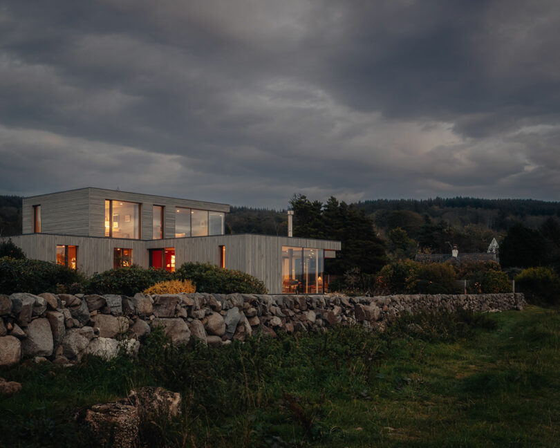 Modern house with large windows and glowing interior lights at dusk, surrounded by greenery and a rocky stone wall, under a cloudy sky.