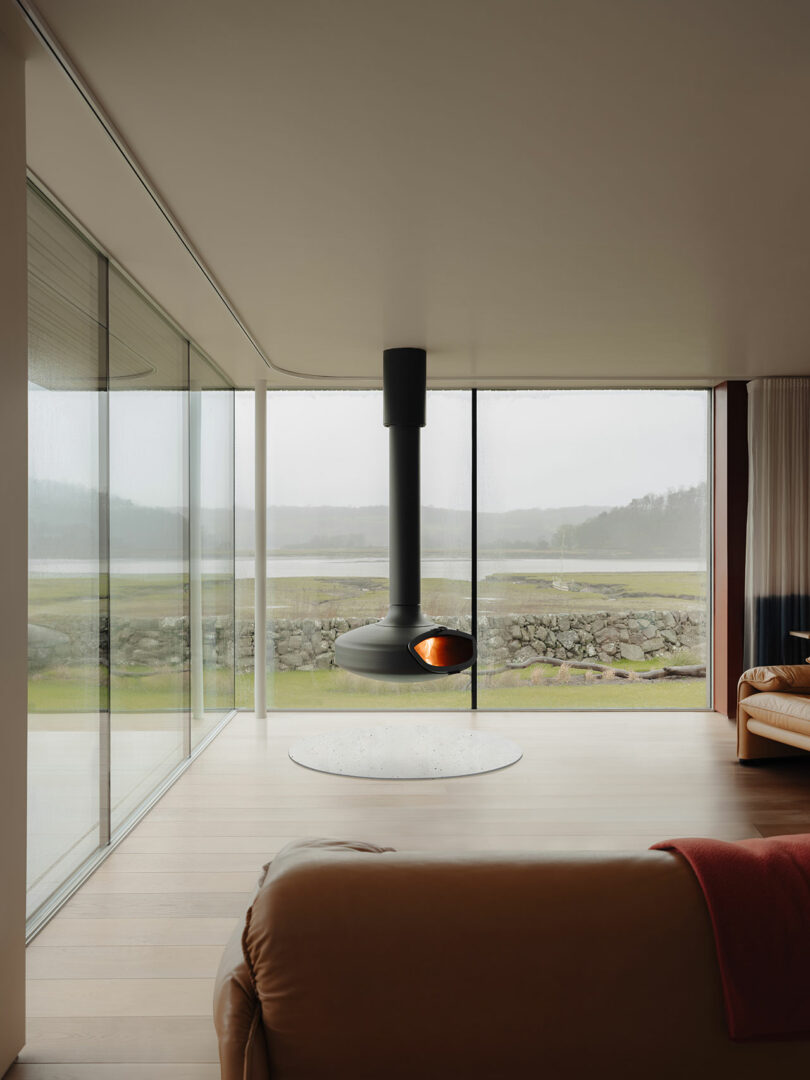 Modern living room with a suspended black fireplace, leather sofas, and large glass windows overlooking a misty landscape.