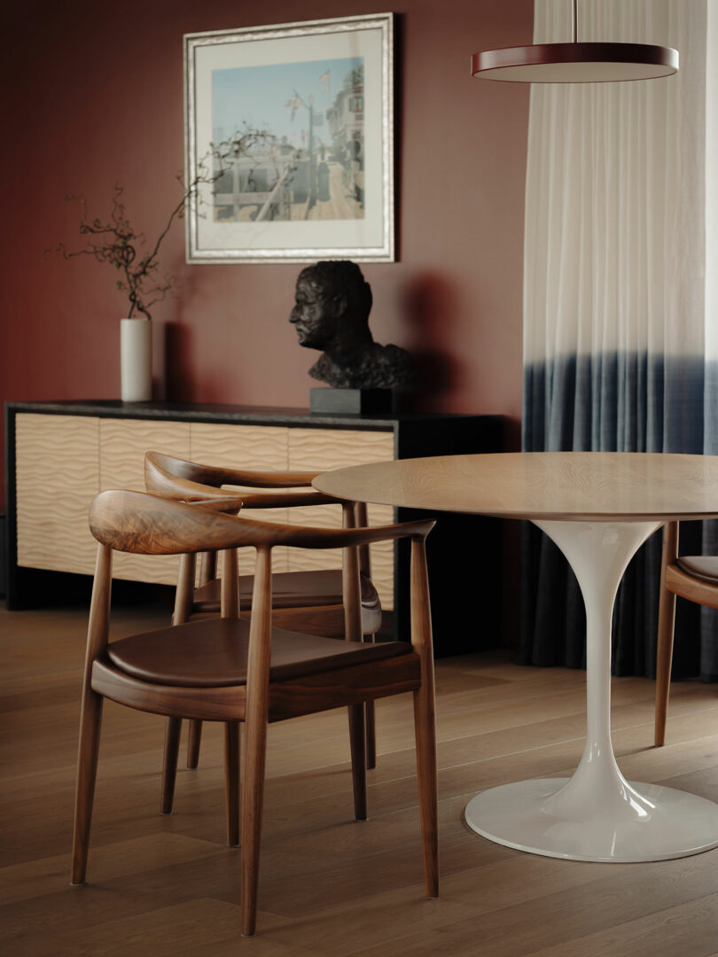 Modern dining room with a round table, wooden chairs, sideboard, and a bust sculpture. A framed painting and vase with branches are on the sideboard. The room has red walls and wooden flooring.