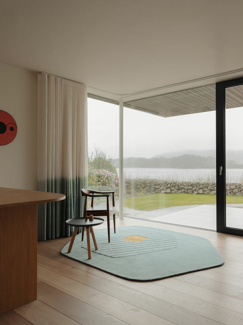 Modern interior with a light wood floor, a wooden chair and table on a green rug. Large windows reveal an outdoor view with a river and trees. A circular wall decoration is to the left.