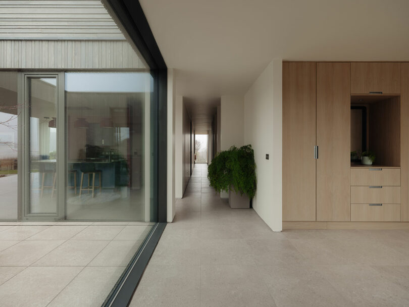 Modern interior with large glass windows, light-colored walls, and tiled floors. A hallway leads to a bright space, with plants and wooden cabinetry visible.