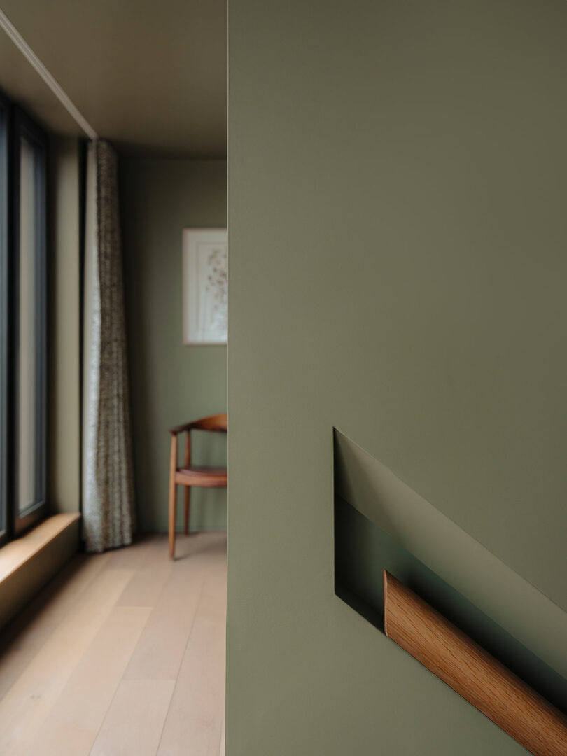 Minimalist interior with green walls, a wooden handrail, and a wooden chair by a window with curtains. A framed picture is on the wall.