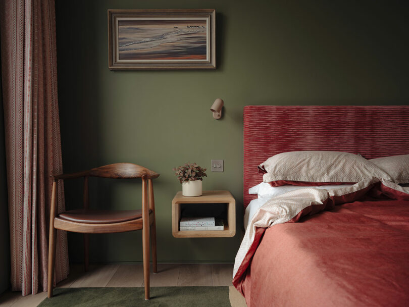 A bedroom with green walls, a red bed, and a wooden chair. A floating shelf holds a plant, and a painting hangs above. Pink curtains are partially visible on the left.