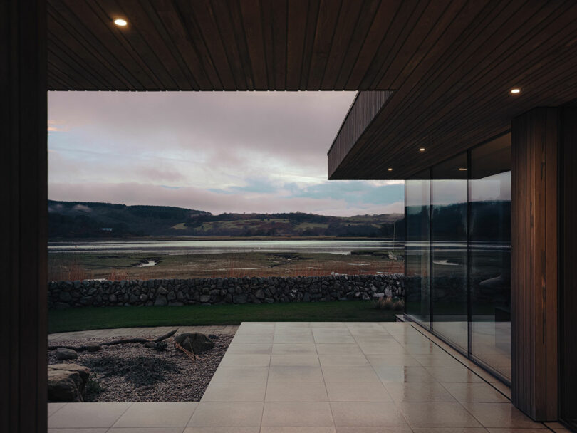 Modern building with a wooden ceiling and glass walls overlooking a scenic view of a lake, stone wall, and hills under a cloudy sky.