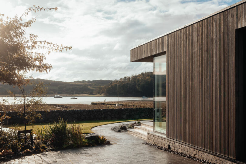 Modern wooden building with large glass window overlooks a serene lakeside view. Pathway leads through landscaped garden. Overcast sky with boats in the distance.