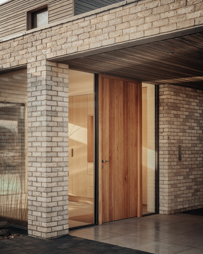 Modern house entrance with large wooden door, surrounded by light brick walls and floor-to-ceiling windows, reflecting warm lighting from the interior.