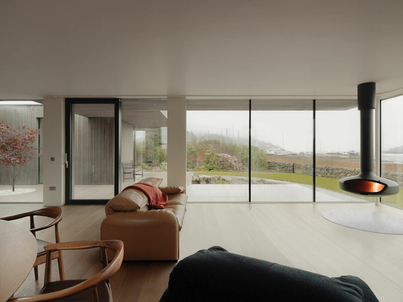 Modern living room with large windows, a beige sofa, wooden chairs, and a suspended fireplace. The view outside shows a misty landscape with greenery.