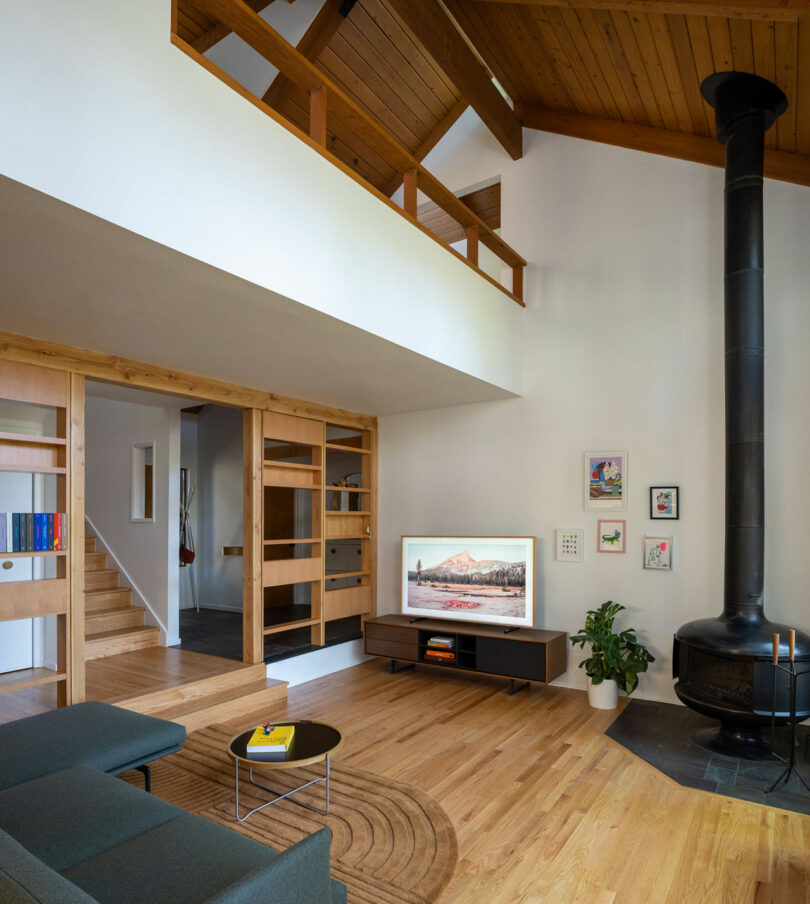 A modern living room with high ceilings, a black suspended fireplace, a flat-screen TV, wooden flooring, a gray sofa, a round coffee table, and a staircase leading to an upper floor.