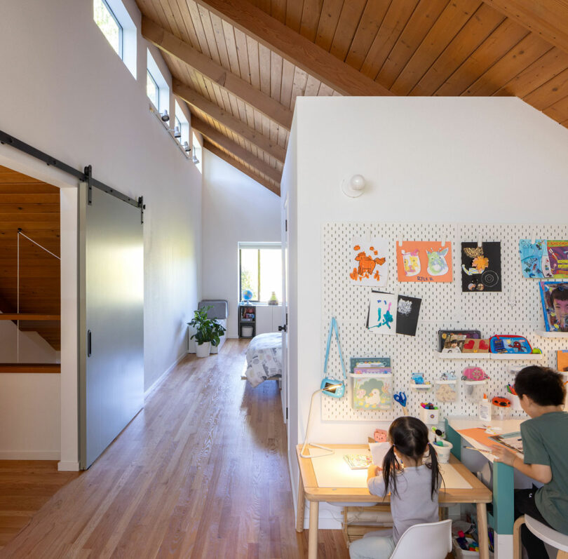 Two children sit at a desk drawing, surrounded by colorful artwork in a bright room with wooden floors and a high, angled ceiling.