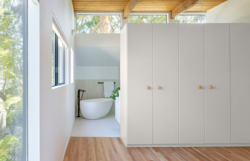 Modern bathroom with a freestanding white bathtub, black faucet, and large windows. Light wood floors and minimalist light gray cabinets are visible. Trees are seen through the windows.