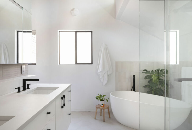 Minimalist bathroom with a white bathtub, glass shower, and black faucet. A small plant sits on a stool next to the tub, and a towel hangs on the wall.